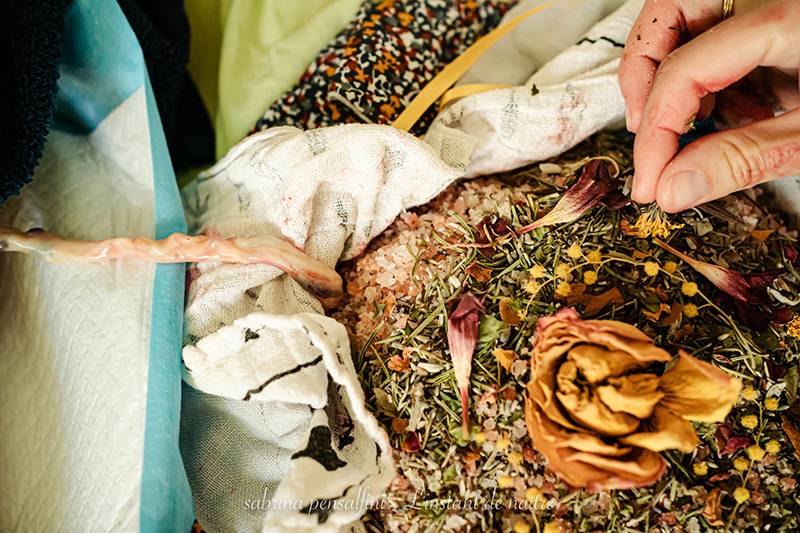 photo d'un placenta lotus lors d'un accouchement à domicile. le placenta est dans un sac en tissus, recouvert de sel et de plantes et fleurs séchées. On voit un bout de cordon ombilical et les doigts d'une femme, la doula, qui installe les plantes séchées. Doula Arles Tarascon